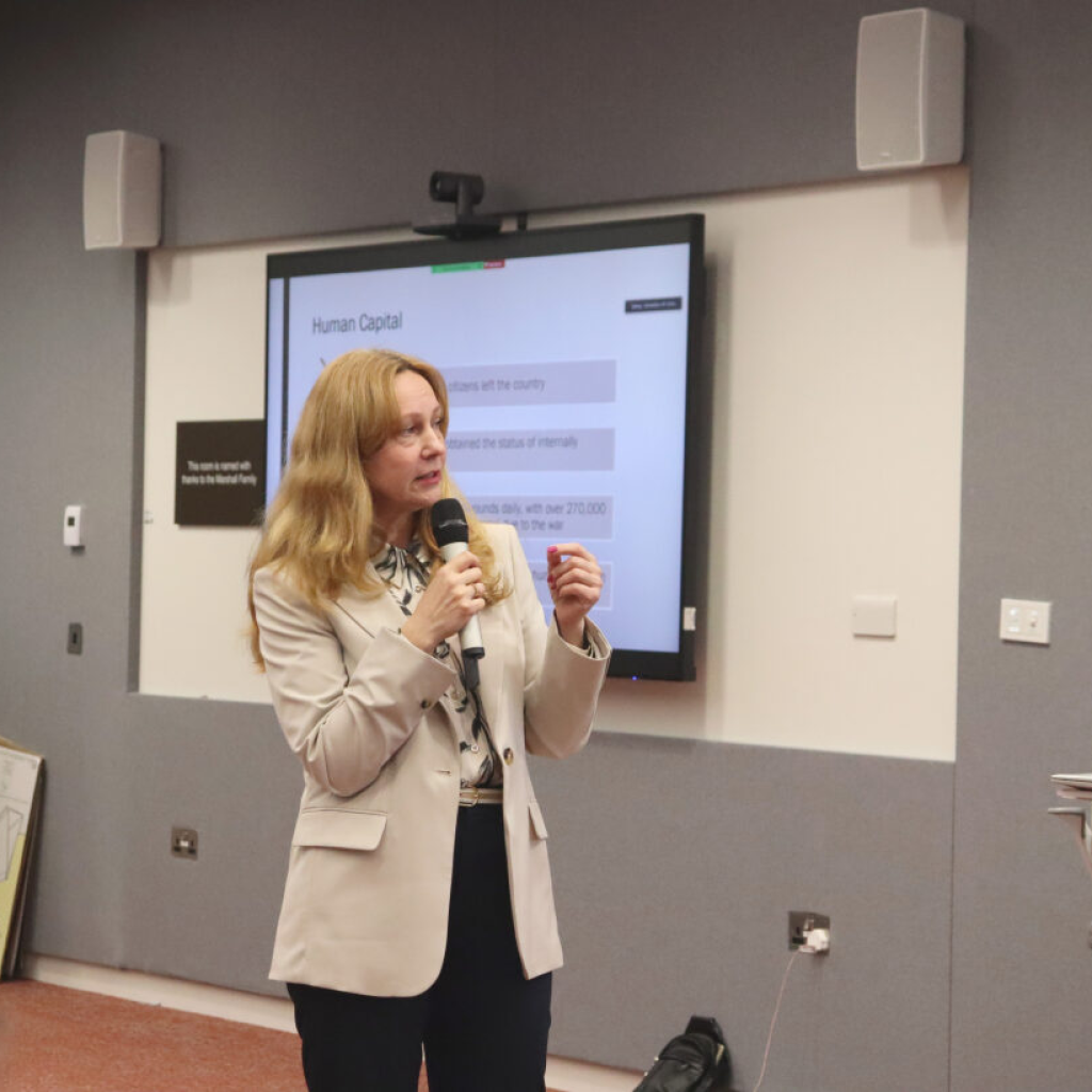 Nataliya Drozd stands in front of presentation slide in a teaching room in the University of Glasgow, speaking into a microphone.